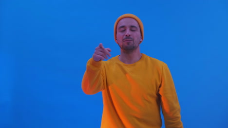 Ein-Mann-Mit-Gelbem-Kapuzenpullover-Und-Hut-Tanzt-Auf-Blauem-Hintergrund.-Lustiges-Tanzen-Beim-Blick-In-Die-Kamera
